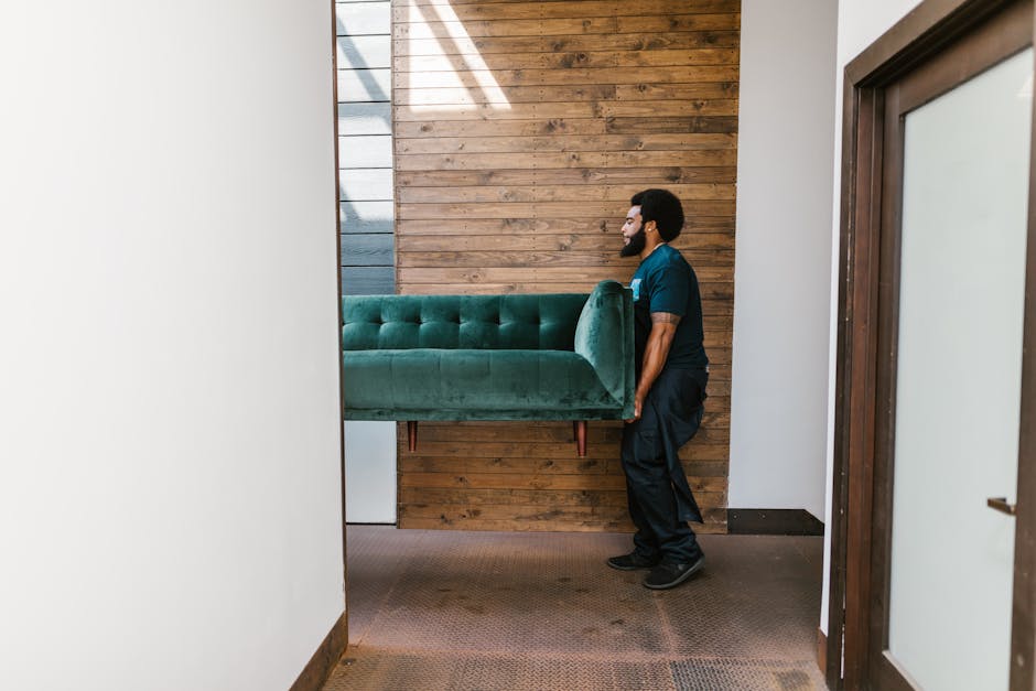A man from Man and Van Palmers Green is engaged in a house removal process, carefully lifting a green upholstered sofa through an interior doorway. The sofa is partially wrapped with packing materials