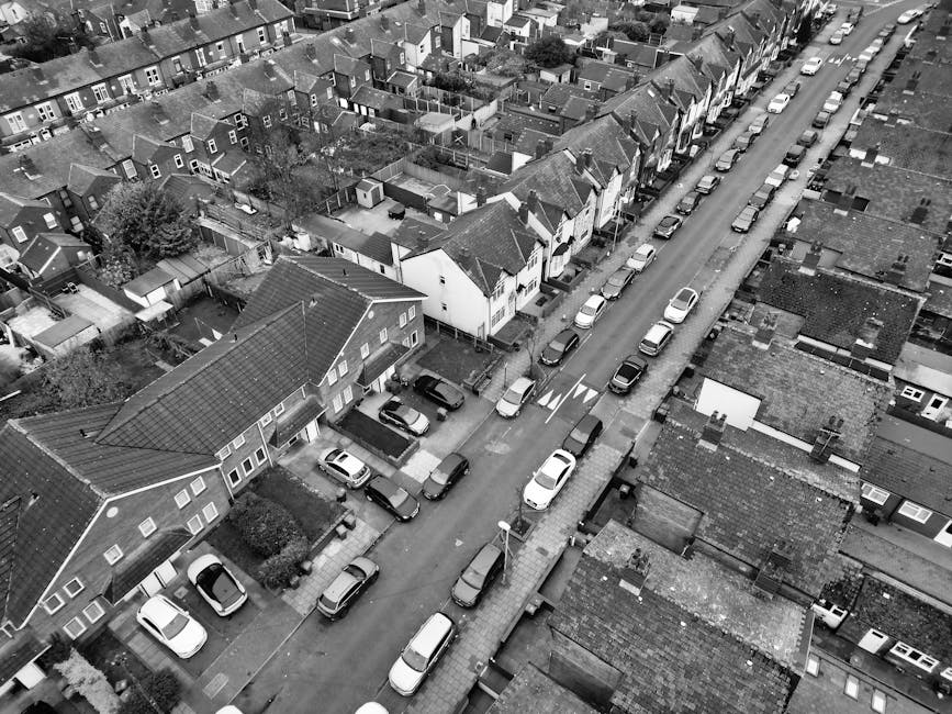 A black-and-white aerial photograph of a narrow residential street in a densely populated area, showing rows of terraced houses with sloped roofs, some with small garden yards, and a variety of parked cars lining both sides of the street. In the foreground, a house with a pitched roof and a small front garden is visible, along with a driveway where a vehicle is parked. Nearby, a man is seen handling cardboard boxes and packing materials, preparing for a home relocation. Next to this, a large van, likely used for furniture transport and moving logistics, is parked on the pavement, with some wrapped furniture or large boxes being loaded or unloaded. The surrounding environment includes other houses, small trees, and utility poles with overhead wires. The scene captures the loading process involved in house removals, illustrating the typical movement of packed belongings, boxes, and furniture during a house clearance or relocation service, as carried out by Man and Van Palmers Green.
