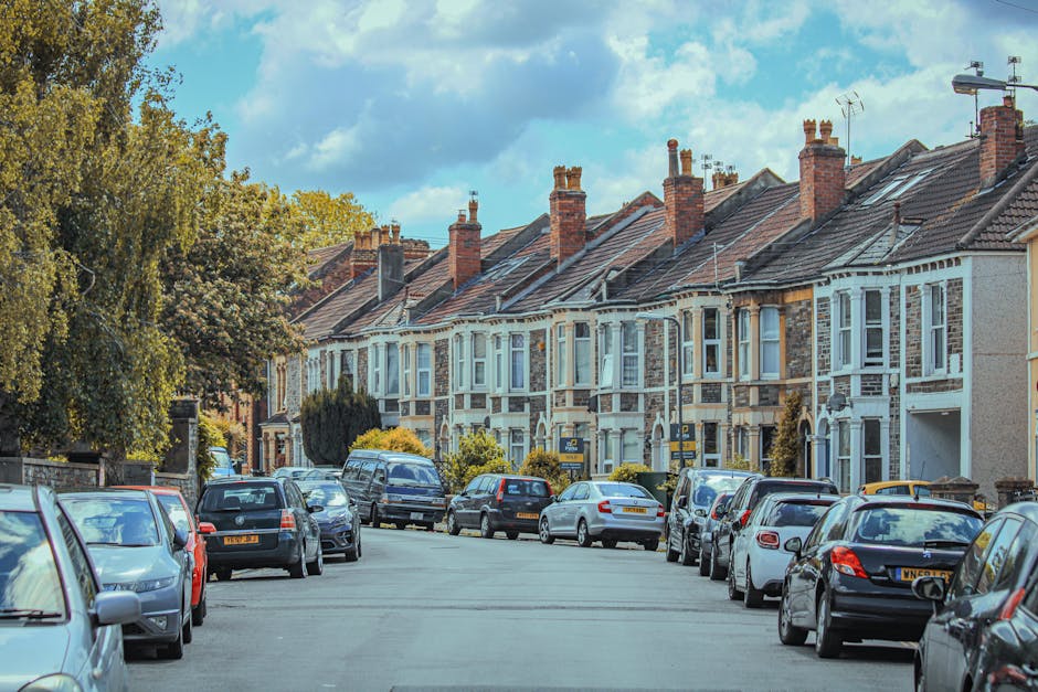 A quiet street lined with terraced houses featuring brick and white-painted facades, with tiled roofs and multiple chimneys. The pavement is occupied by parked cars of various makes and models, parked parallel to the kerb. The street is bordered by trees with green foliage, and the sky above shows a mix of clouds and blue patches. In the context of home relocation services, this image illustrates a typical residential area where furniture and boxes might be transported during a house move, with visible vehicles and the street environment indicating potential loading and moving logistics handled by Man and Van Palmers Green. The scene captures the outdoor environment suitable for loading furniture onto a van for a comprehensive removals process, emphasizing the importance of planning and careful packing in narrow street settings for terraced houses.