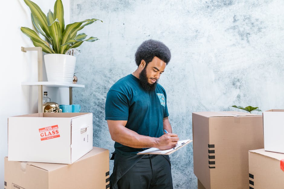 A professional mover from Man and Van Palmers Green is standing inside a room with light blue textured walls, surrounded by packed cardboard boxes for a house or office relocation. He is holding a clipboard and pen, carefully noting the contents of the boxes, which include a large box marked 'GLASS' in red tape, a cardboard box with black stripes, and other sealed cartons. A white shelf behind him holds a potted plant with broad green leaves, decorative objects, and a few small storage containers. The scene shows the loading process of furniture transport and packing for a home or business move, with natural indoor lighting illuminating the area and highlighting the careful handling of items in a professional moving environment associated with relocation services.