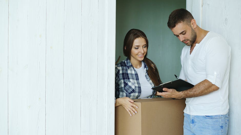 A man and woman with light skin are involved in a house relocation process indoors, with the woman standing behind a partially visible cardboard moving box positioned on the floor, smiling and wearing casual clothing including a checked shirt. The man, dressed in a white t-shirt and jeans, is standing nearby, holding a clipboard and pen, appearing to review or check details related to the packing and moving process. The environment includes a white painted wooden wall and a doorway leading to another room, with natural lighting illuminating the scene, depicting the typical steps involved in furniture transport and packing for a house or office removal service. This image reflects the careful planning and coordination necessary in professional relocation tasks and is relevant to services offered by companies like Man and Van Palmers Green, specialising in removals and business relocations.