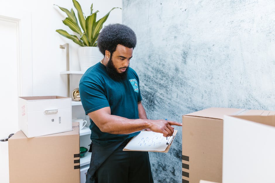A professional mover from Man and Van Palmers Green is standing inside a room with light blue textured walls, surrounded by packed cardboard boxes for a house or office relocation. He is holding a clipboard and pen, carefully noting the contents of the boxes, which include a large box marked 'GLASS' in red tape, a cardboard box with black stripes, and other sealed cartons. A white shelf behind him holds a potted plant with broad green leaves, decorative objects, and a few small storage containers. The scene shows the loading process of furniture transport and packing for a home or business move, with natural indoor lighting illuminating the area and highlighting the careful handling of items in a professional moving environment associated with relocation services.