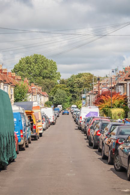 A residential street scene with parked vehicles lining both sides of the narrow tarmac road, including various cars and moving vans, some of which are partially covered with protective tarps. The street is bordered by row houses with brick facades, small front gardens, and trees with lush green foliage, indicating a suburban area. Overhead, multiple power lines crisscross the sky, which is partly cloudy with patches of blue. In the background, larger trees and more houses are visible, suggesting a quiet neighbourhood. The image captures a typical urban environment suitable for home relocation or furniture transport, often involved in house removals services like those provided by Man and Van Palmers Green, aligning with the concept of packing and moving logistics in residential areas.