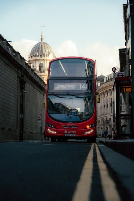 A red double-decker bus with a large front windshield and the route number 14, branded with the Palmers Green destination, is parked on a narrow street between tall stone or concrete buildings in an urban area. The bus is part of the Arriva fleet and is positioned head-on in the image, with the lower front facing the camera, occupying the central part of the scene. The street features yellow double line markings along the curb, indicating parking or stopping restrictions, and extends into the background where other vehicles and pedestrians may be present. On the left, a building with a domed roof topped with a spire is visible beyond the bus, while on the right, a bus stop shelter with a timetable display and adjacent signage is evident. The sky above is partly cloudy, with daylight illuminating the scene. This image captures the typical environment for house removals and furniture transport within the city, exemplifying moving logistics involving public transport routes and urban infrastructure, as related to professional removals services such as those offered by Man and Van Palmers Green.