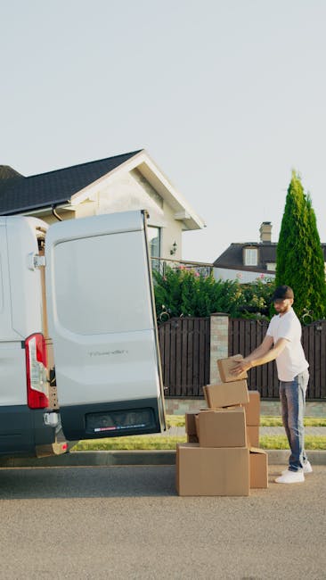 A man wearing a black shirt and blue jeans is pushing a trolley loaded with large cardboard boxes and wrapped furniture, possibly mattresses, through an outdoor urban area beneath an overpass or bridg