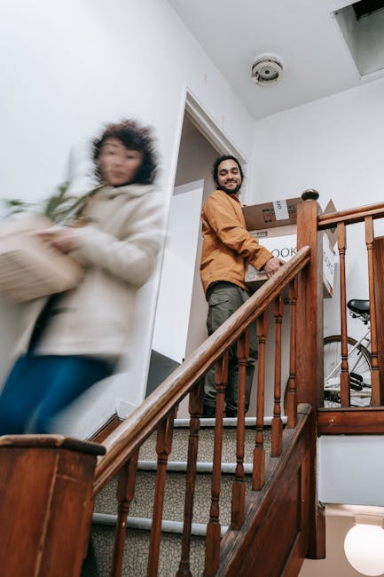 Inside a residential property, a man with a beard and wearing an orange jacket is standing on a carpeted staircase, engaged in the home relocation process by holding moving boxes. The staircase features wooden balustrades and newel posts, leading up from a lower level where a bicycle is partially visible. An open doorway behind the man reveals a room with white walls and a ceiling-mounted smoke detector. To the left, a woman dressed in a cream-colored coat and blue jeans appears to be moving quickly past the scene, holding a cardboard box wrapped in green packing paper, causing her motion blur. The environment is lit by natural light, and the scene captures a typical furniture transport or packing and moving situation, with attention to spatial arrangement, materials like cardboard and plastic wrap, and moving equipment visible. This image exemplifies the logistical aspects of house removals and stair access solutions, as handled by Man and Van Palmers Green, supporting smooth removal services.
