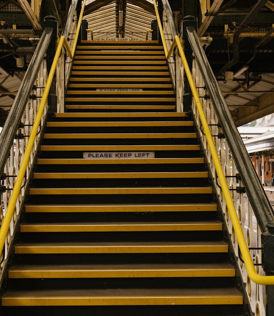 An industrial staircase with yellow safety handrails on both sides and black anti-slip treads leads upward to an enclosed upper level. The staircase is situated inside a building with exposed metal beams and piping visible overhead. Attached to the middle of the lower steps is a white sign with black text stating, 'PLEASE KEEP LEFT.' The environment appears to be a warehouse or storage area used for house removals or furniture transport, with a focus on safe movement and loading of large items. The lighting is industrial, casting even illumination across the stairs, which are part of a loading or access point for moving furniture or packing materials during a house relocation conducted by Man and Van Palmers Green.