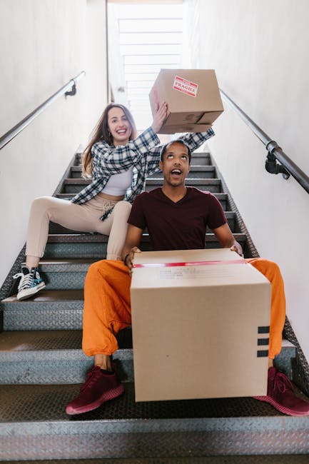 Inside a residential property, a man with a beard and wearing an orange jacket is standing on a carpeted staircase, engaged in the home relocation process by holding moving boxes. The staircase features wooden balustrades and newel posts, leading up from a lower level where a bicycle is partially visible. An open doorway behind the man reveals a room with white walls and a ceiling-mounted smoke detector. To the left, a woman dressed in a cream-colored coat and blue jeans appears to be moving quickly past the scene, holding a cardboard box wrapped in green packing paper, causing her motion blur. The environment is lit by natural light, and the scene captures a typical furniture transport or packing and moving situation, with attention to spatial arrangement, materials like cardboard and plastic wrap, and moving equipment visible. This image exemplifies the logistical aspects of house removals and stair access solutions, as handled by Man and Van Palmers Green, supporting smooth removal services.