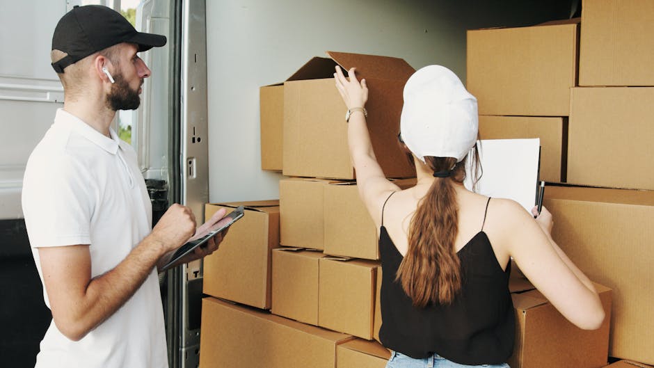 A man and a woman are standing next to a stack of cardboard moving boxes inside a van or storage area, which is part of a home relocation process. The woman, wearing a white cap and black sleeveless top, is reaching up to adjust or inspect a box on top of the stack, holding a clipboard in her other hand. The man, dressed in a white shirt and black cap, is holding a tablet or clipboard and appears to be recording or checking information. The boxes vary in size and are arranged in an organized manner against the interior wall of the vehicle. Foam padding or packing materials are visible around some of the boxes to protect fragile items during furniture transport. The environment suggests a loading or packing activity associated with professional removals services, as provided by Man and Van Palmers Green, supporting efficient packing and moving processes within an interior property or vehicle loading area.