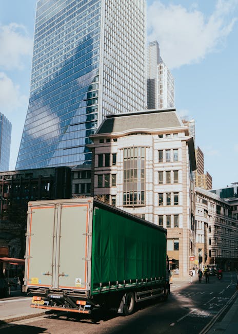 A large green moving truck is parked on a city street in front of a mix of modern glass skyscrapers and older stone buildings. The truck appears to be involved in a home relocation or furniture transport process, with its back doors closed. The street has some pedestrians and a few vehicles parked nearby, under a clear blue sky with a few clouds. The scene captures the typical urban environment associated with house removals and professional moving services, with the truck ready for loading or unloading of household furniture, boxes, or appliances as part of a scheduled move. Man and Van Palmers Green operates in this area, offering removals and packing logistics for residential relocations.