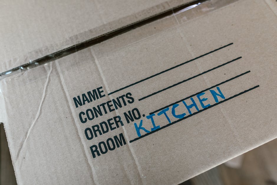 A man and a woman are standing next to a stack of cardboard moving boxes inside a van or storage area, which is part of a home relocation process. The woman, wearing a white cap and black sleeveless top, is reaching up to adjust or inspect a box on top of the stack, holding a clipboard in her other hand. The man, dressed in a white shirt and black cap, is holding a tablet or clipboard and appears to be recording or checking information. The boxes vary in size and are arranged in an organized manner against the interior wall of the vehicle. Foam padding or packing materials are visible around some of the boxes to protect fragile items during furniture transport. The environment suggests a loading or packing activity associated with professional removals services, as provided by Man and Van Palmers Green, supporting efficient packing and moving processes within an interior property or vehicle loading area.