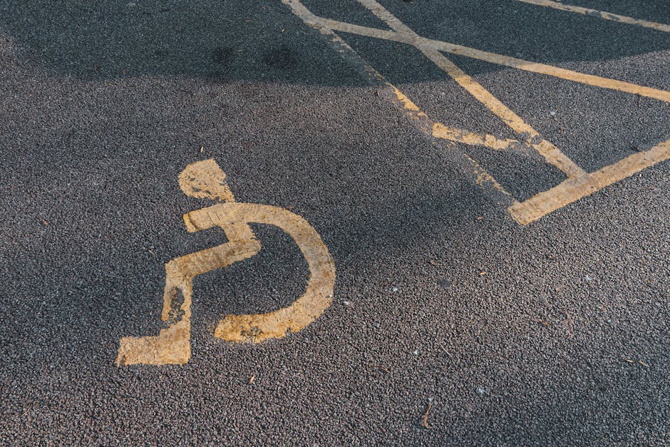 A close-up of a designated accessible parking space on a tarmac surface, featuring a yellow-painted wheelchair symbol. The paint is slightly worn, showing some scratches and fading. The parking space 